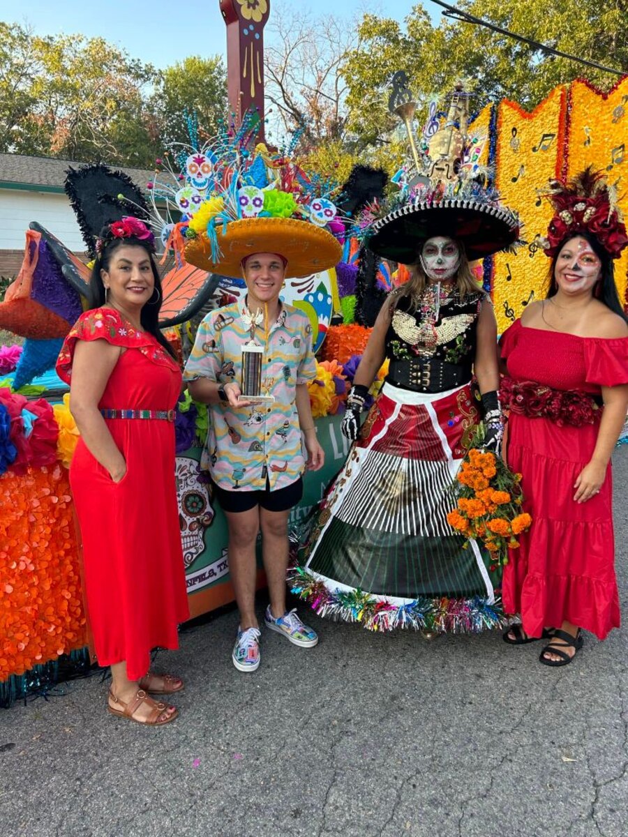 Festival volunteers and a Dia de los Muertos parade float winner posing with an award trophy in front of a colorful decorated float