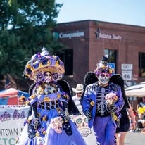 A couple in matching purple Dia de los Muertos costumes and skeleton face paint walking in the procession parade