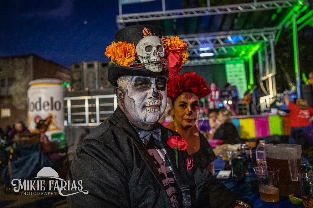 A couple in Dia de los Muertos skeleton face paint, the man in a marigold-and-skull top hat, smiling together at the festival