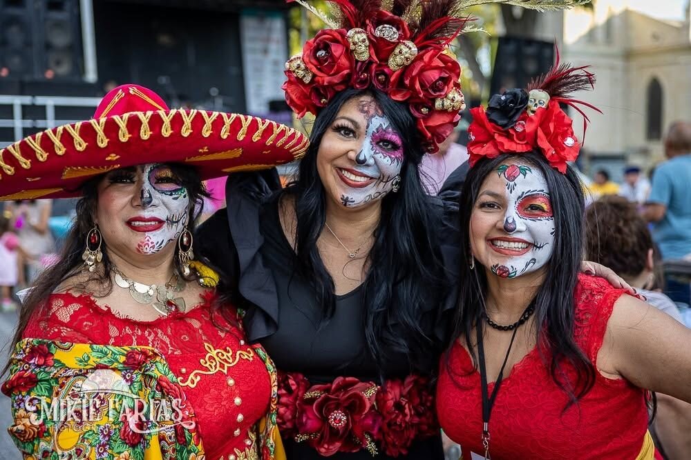 Three women in vibrant Dia de los Muertos face paint and flower crowns, one wearing a red sombrero, smiling together at the festival
