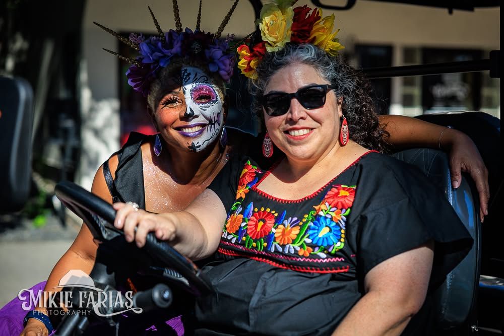 Two friends smiling together in a golf cart at the festival, one in elaborate Dia de los Muertos sugar skull face paint and a floral crown, the other in a traditional embroidered dress and sunglasses with a marigold flower crown