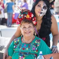 Two women celebrating together, one in a green embroidered dress, at the Dia de los Muertos festival