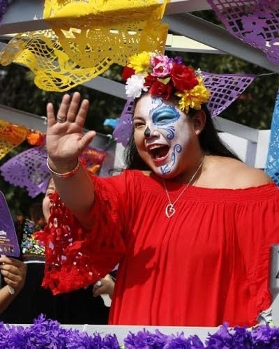Tanya Castilleja Perez smiling and waving, wearing a red off-shoulder blouse and Dia de los Muertos sugar skull face paint with a colorful flower crown, at the festival parade
